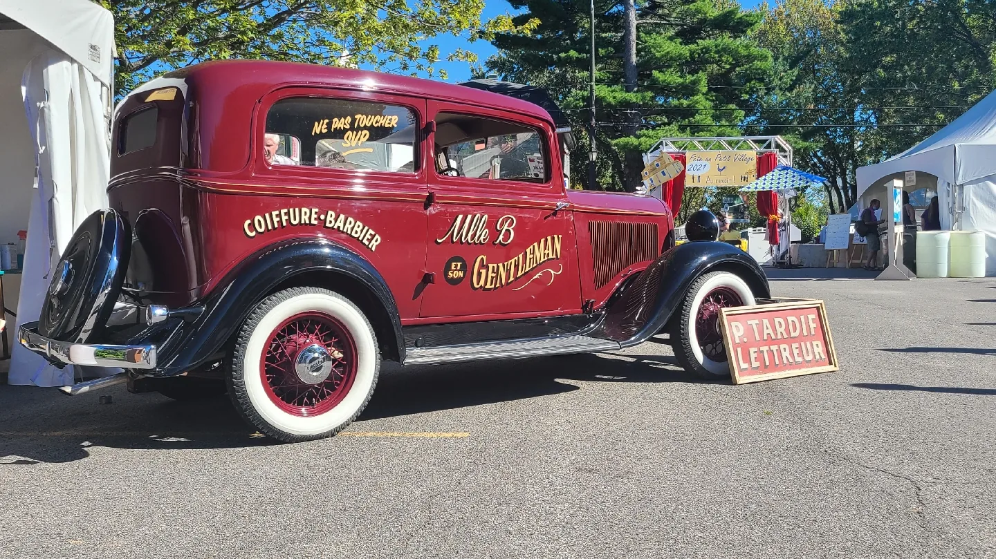 Une automobile ancienne bordeaux aux roues à rayons et pneus à flancs blancs, lettrée à la main en doré : « Coiffure · Barbier » et « Mlle B et son Gentleman ».